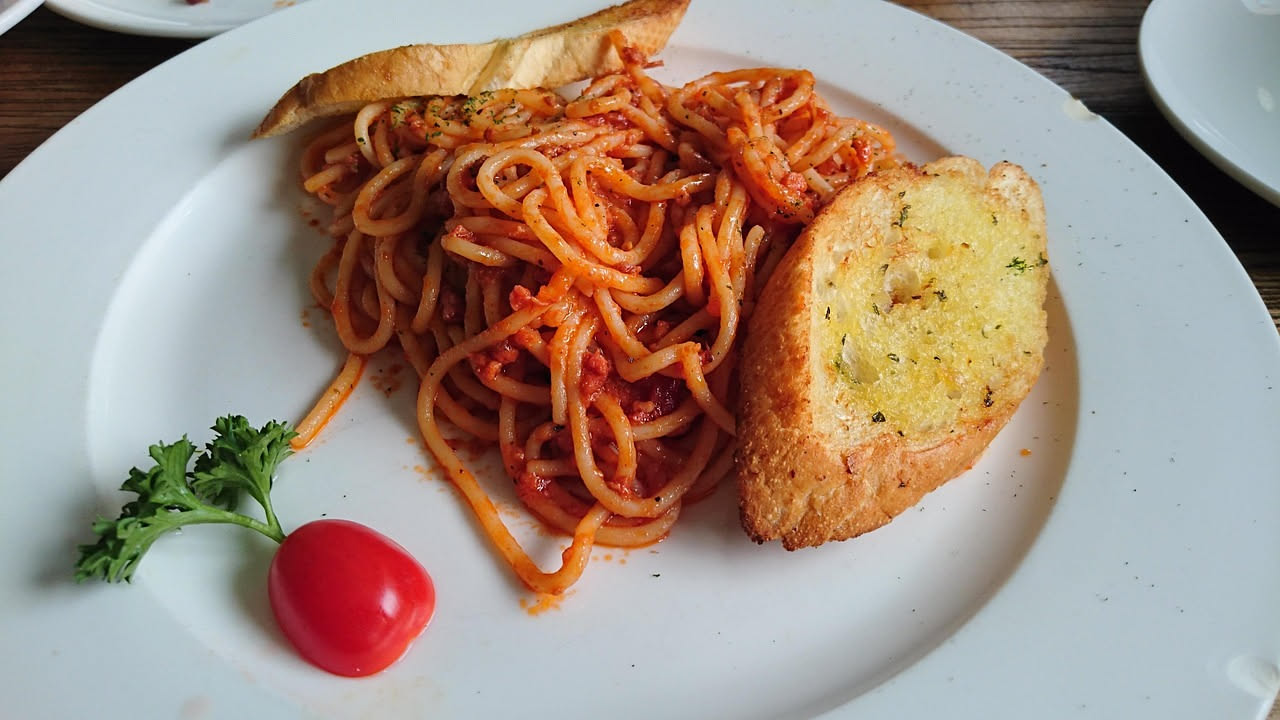 Un piatto di spaghetti conditi con salsa rossa densa, guanciale croccante in piccoli cubetti e una spolverata di pecorino grattugiato giallastro, con basilico fresco verde sul bordo bianco del piatto