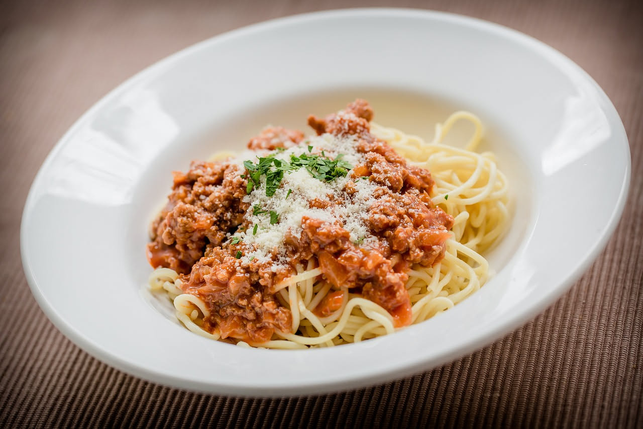 Piatto bianco con spaghetti gialli ricoperti di ragù rosso scuro con particelle di carne visibile, grattugia di parmigiano bianca in superficie, forchetta sul bordo