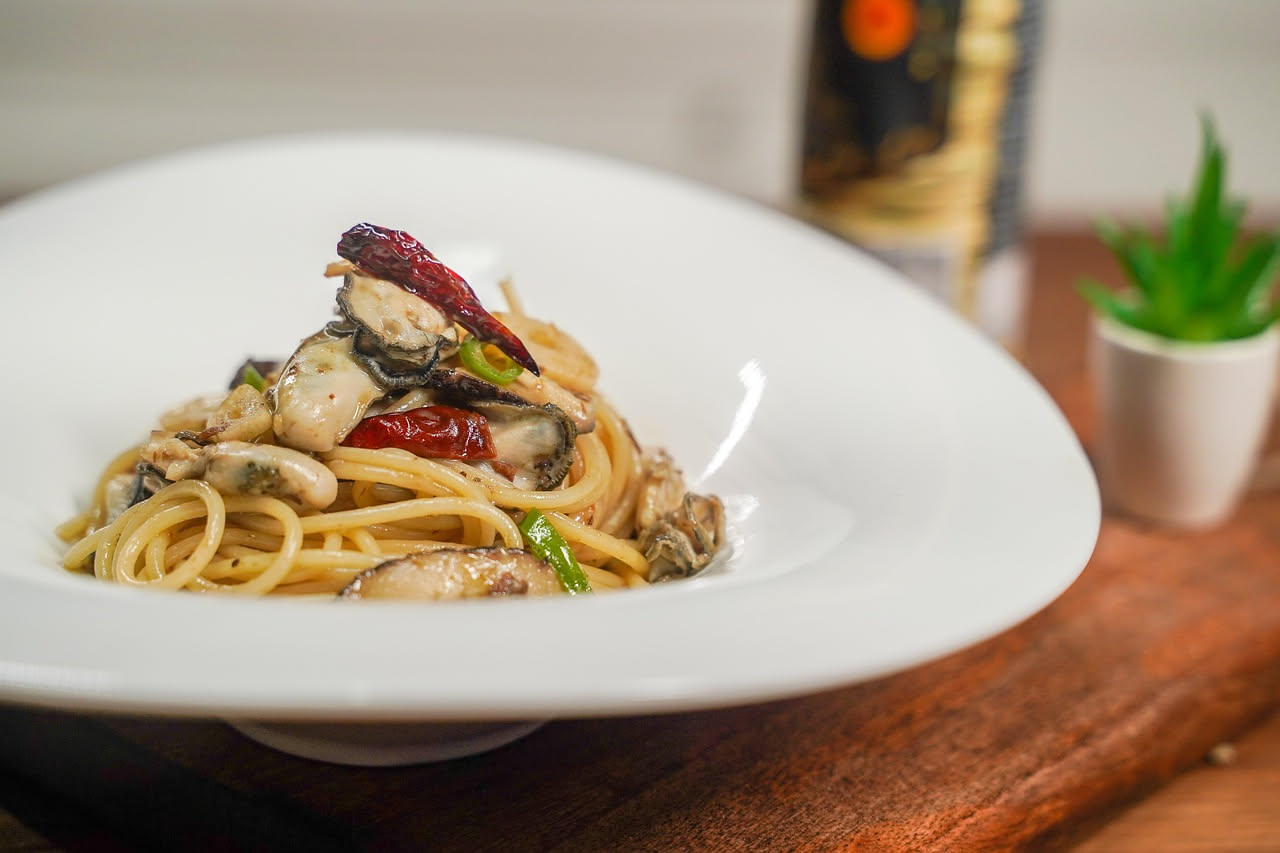 Un piatto di porcellana bianca con spaghetti al dente bianchi e beige, distribuiti uniformemente, con ostriche aperte grigiastre sparse sopra, perlato lucido delle valve, aglio dorato e prezzemolo ver