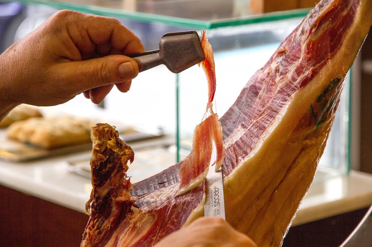 Speck rosa pallido affettato finissimo, leggermente arricciato, disposto in cerchio su piatto bianco con fette di pane tostato ai margini e rucola fresca