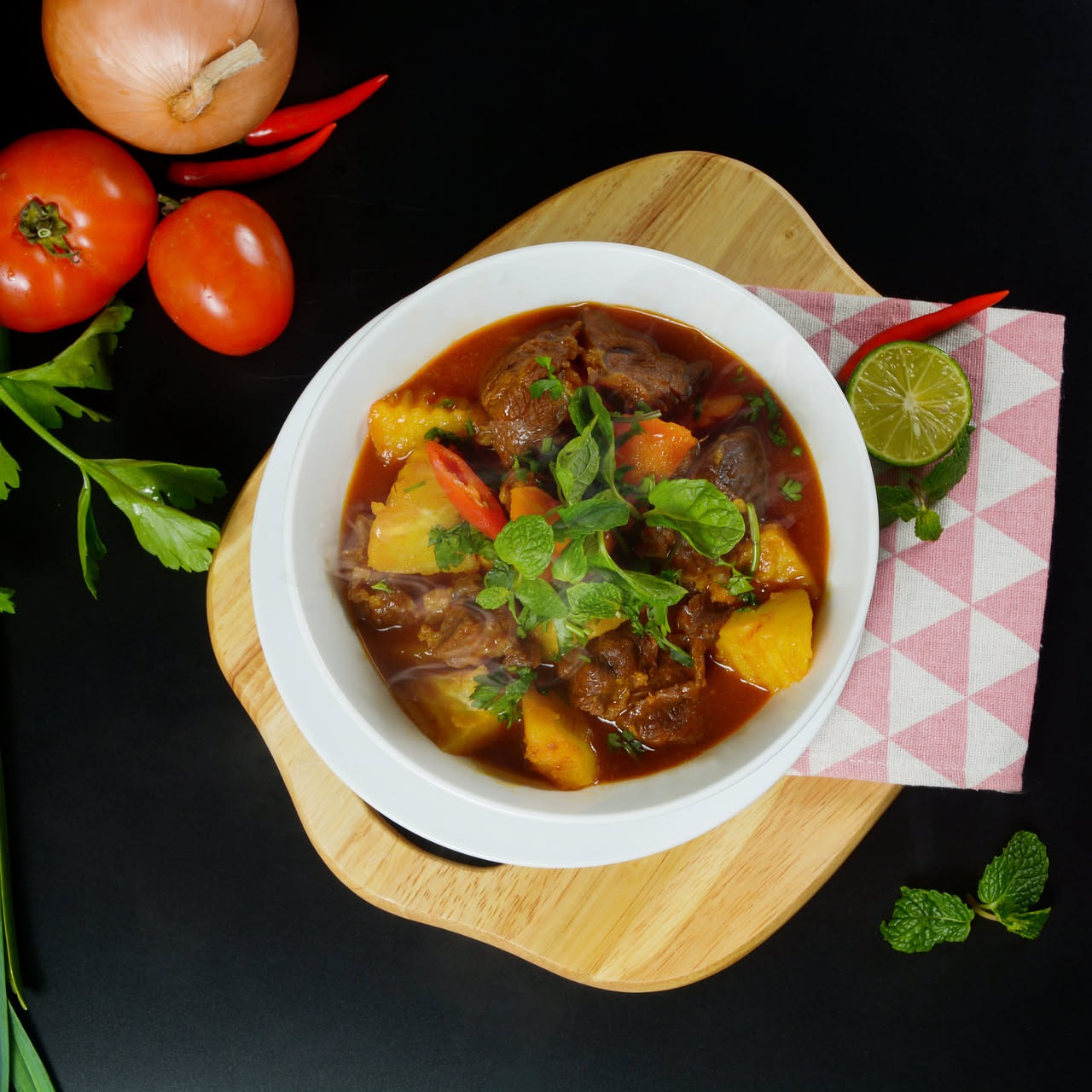 Spezzatino di capra rosso mattone in un piatto di ceramica bianca, pezzi di carne morbida in sugo denso, con carote arancioni e patate biancastre, cipolle scure visibili, guarnito con rosmarino fresco