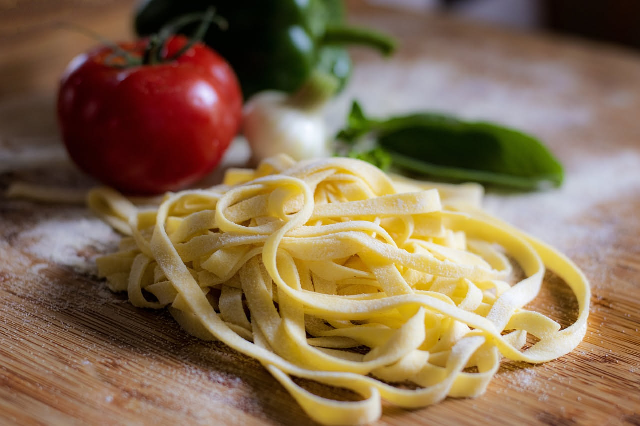 Piatto bianco con tagliatelle dorate ricoperte di sugo scuro e denso, guarnite con parmigiano grattugiato e una foglia di salvia, su tavolo di legno chiaro con bicchiere di vino rosso sfocato sullo sf