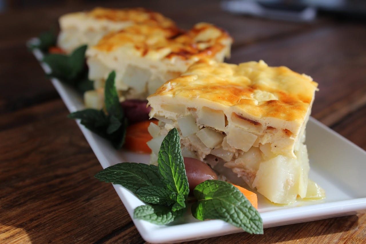 Timballo teramano intero appena sfornato con pasta sfoglia dorata, in piedi dentro una teglia da forno, con vapore leggero che sale dalla superficie; sullo sfondo un piatto bianco con una porzione ser