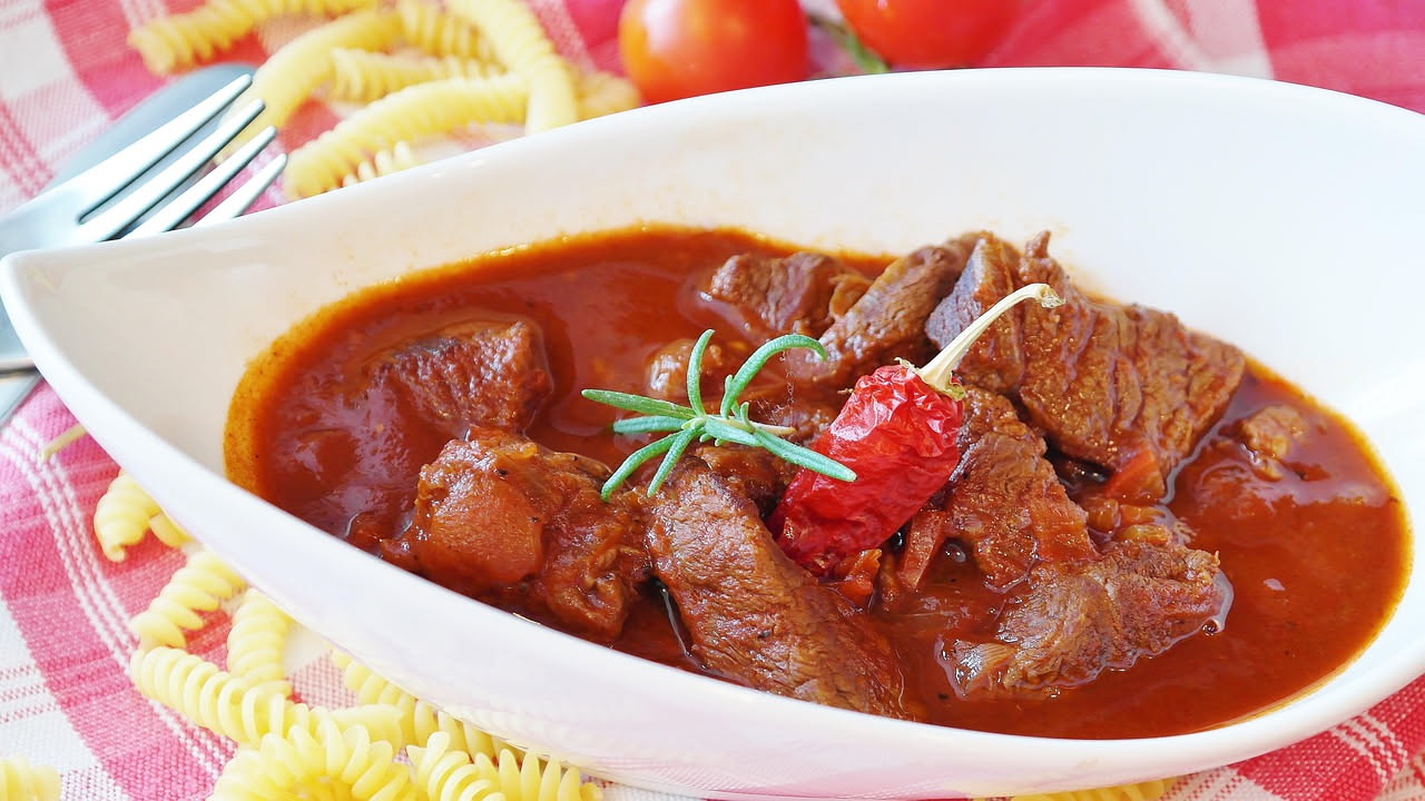 Trippa rosso scuro in umido in una ciotola di ceramica bianca, con pomodori rossi e peperoncino rosso accanto, su tavolo di legno