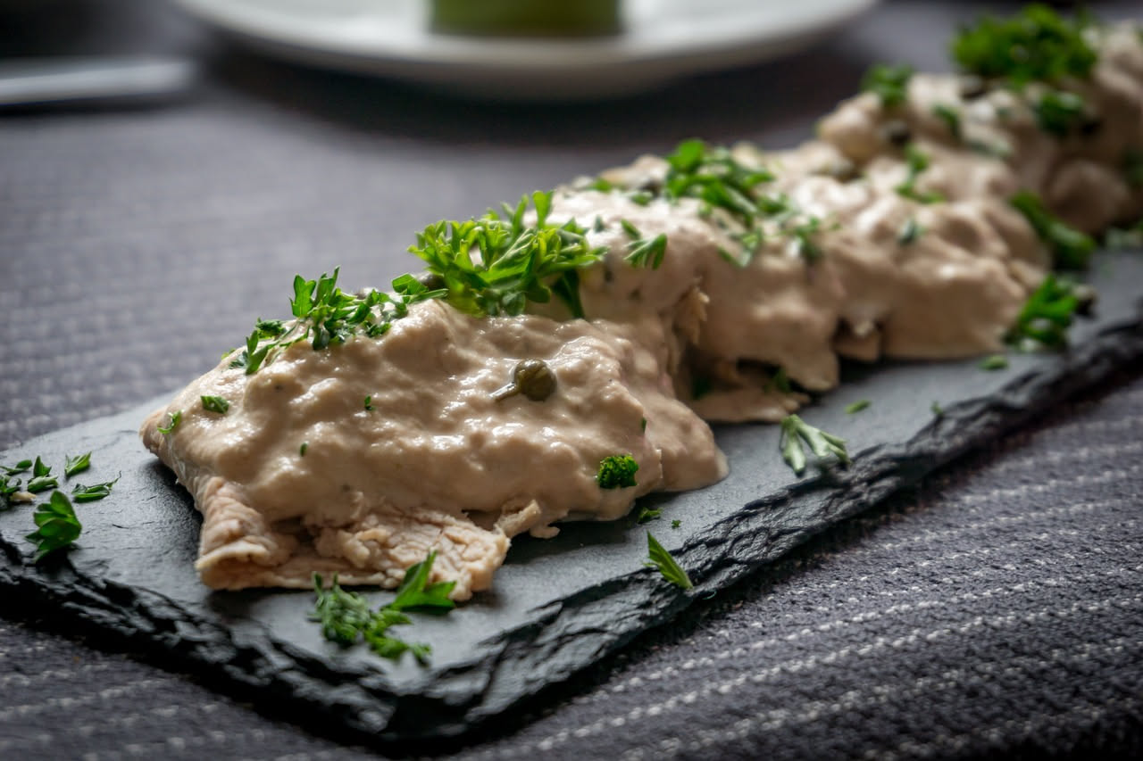 Fette di vitello lesso coperte da salsa tonnata densa con striature bianche e grigie, guarnite con capperi e acciughe, presentate su un piatto di ceramica bianca