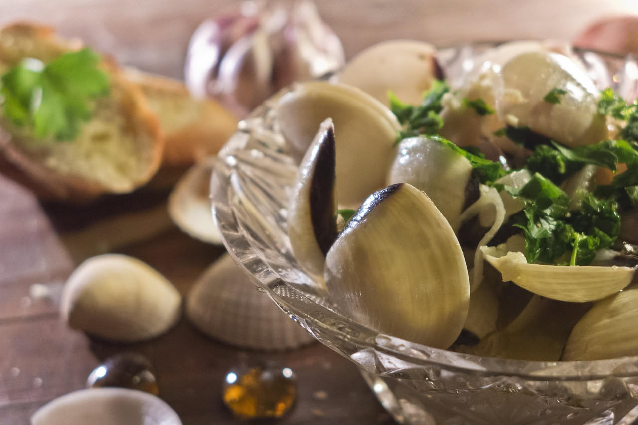 Tre vongole veraci aperte su un piatto bianco con mezzo limone e gocce d'acqua, servite su un tavolo di legno con una bottiglia di olio d'oliva sullo sfondo
