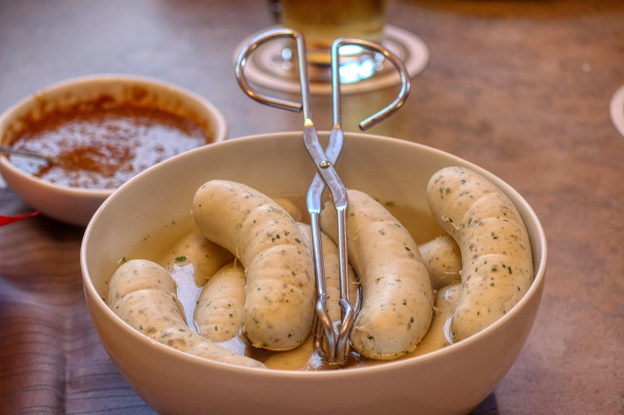 Due Würstel di Bolzano dorati e affumicati disposti in un piatto bianco accanto a senape piccante e pane di segale tostato, con una tazza di brodo fumante sullo sfondo