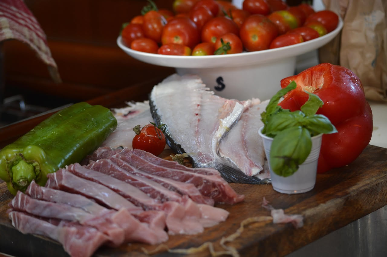 Una ciotola bianca di ceramica con zuppa di pesce fredda: brodo trasparente color ambra con pezzi di pesce bianco, pomodori rossi, sedano verde, e fettine di limone galleggianti. Accanto, una fetta di