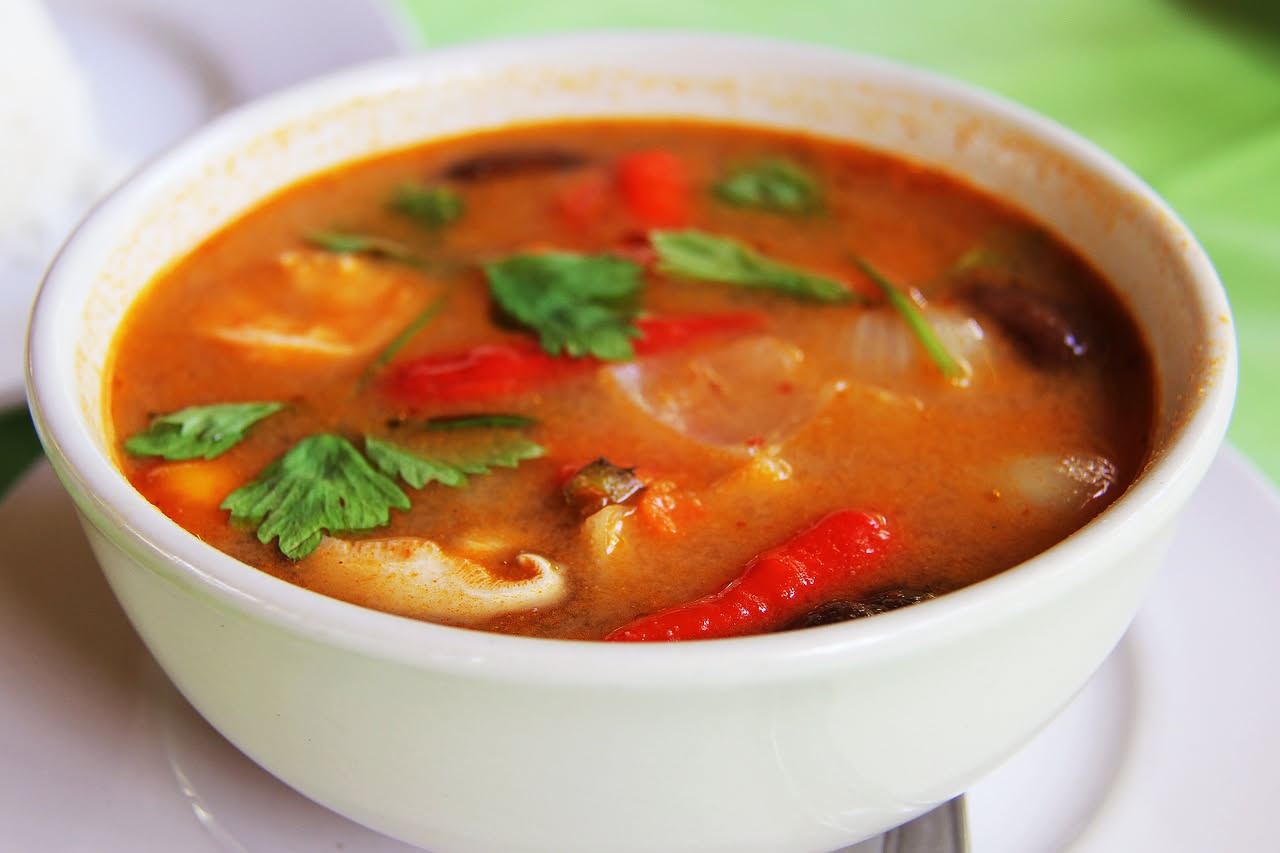 Zuppa di pesce in ciotola di ceramica bianca: brodo trasparente con vongole aperte, gamberi rosa, filetti di pesce bianco, pomodori rossi, prezzemolo fresco verde, servita con pane tostato ai lati