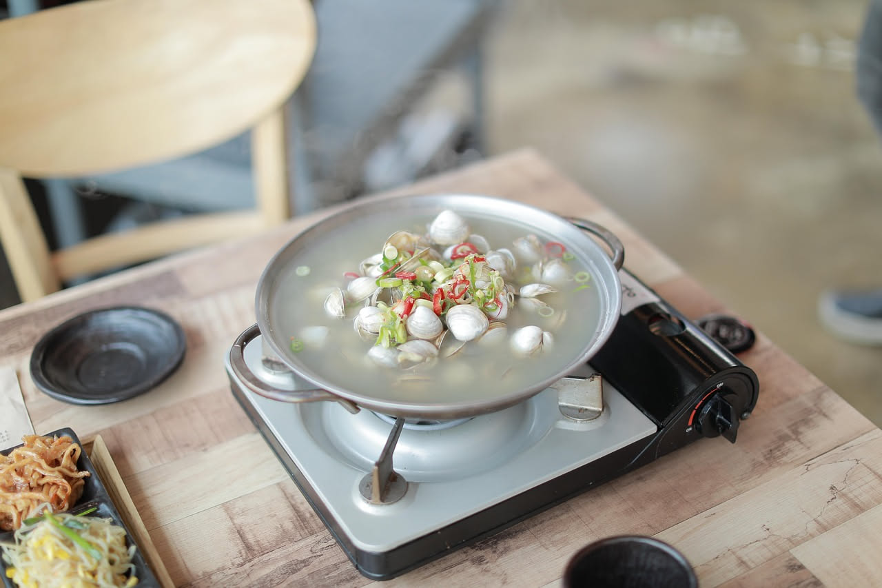 Zuppa di vongole veraci in ciotola bianca fonda: vongole aperte e piene di carne biancastra dentro guscio grigio scuro, immerse in brodo trasparente giallo-arancio, aglio soffritto visibile, prezzemol
