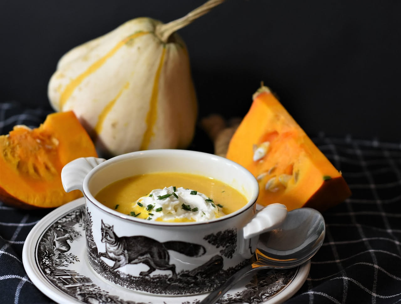 Una ciotola bianca di zuppa di zucca cremosa color arancione, guarnita con semi di zucca tostati e fili di olio verde, con una cucchiaiata sul bordo, vista dall'alto su tavolo di legno grigio