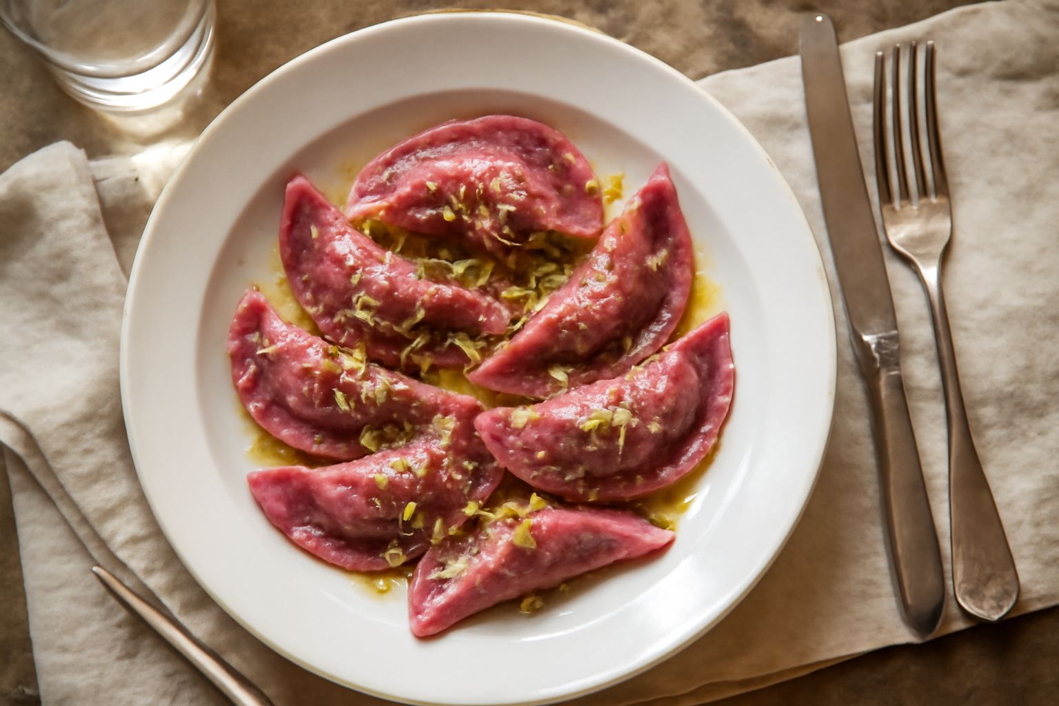Casunziei bergamaschi cotti su piatto bianco, di colore dorato, a forma di mezzaluna, guarniti con burro fuso e salvia fresca, accompagnati da una ciotola di parmigiano grattugiato e un bicchiere di v