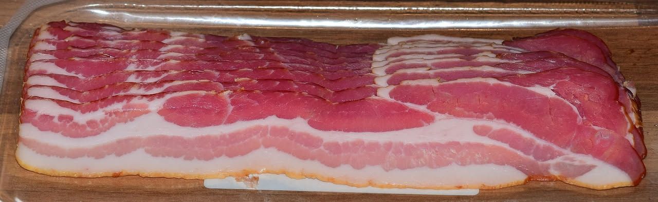 Fette di guanciale modenese affettate sottili su un tagliere di legno, rosso intenso con marezzature bianche di grasso, accanto a un coltello e un bicchiere di vino rosso