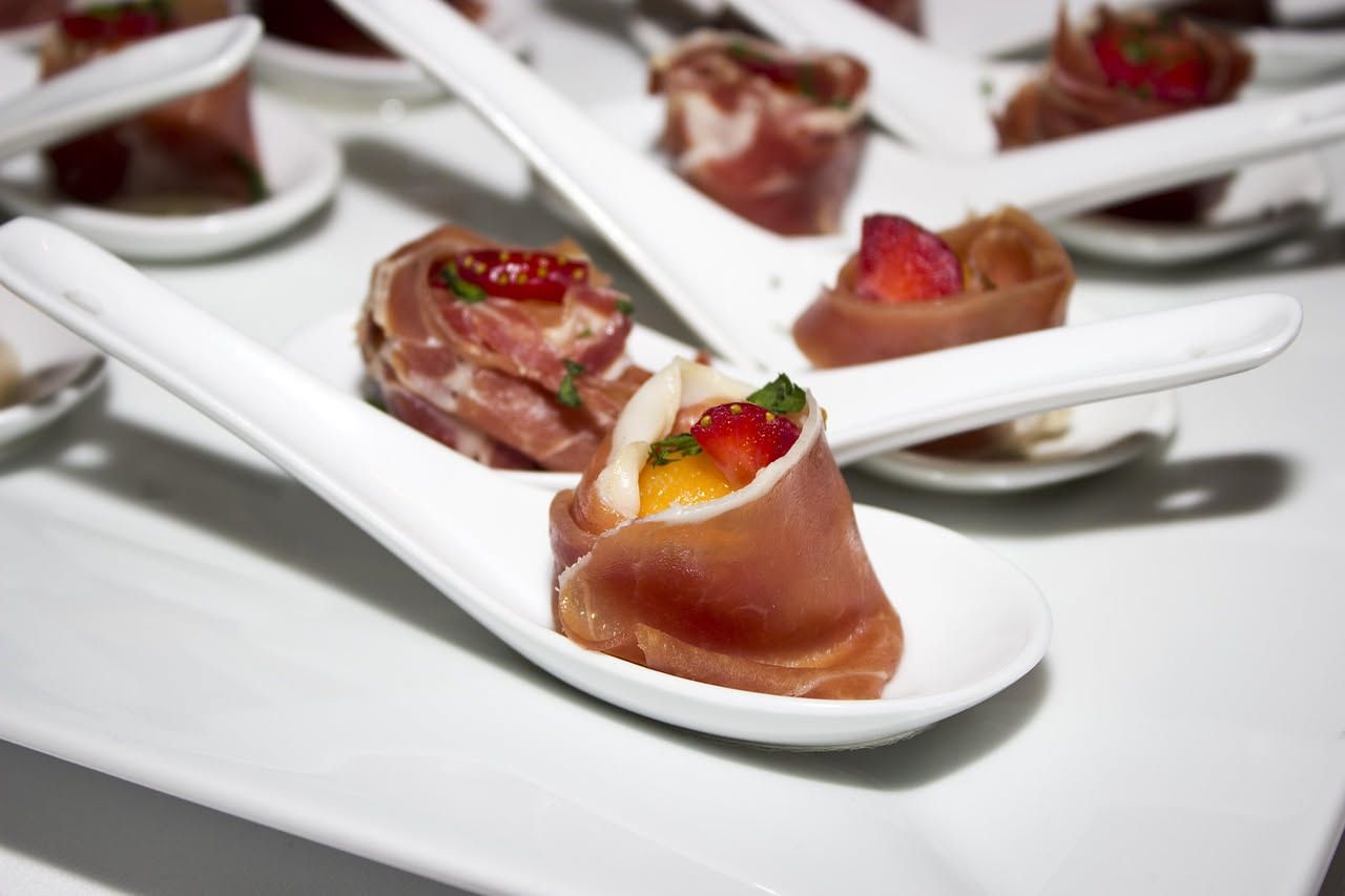 Fette di melone giallo pallido disposte a ventaglio con prosciutto crudo rosato e ricotta salata bianca su un piatto bianco, con basilico fresco verde